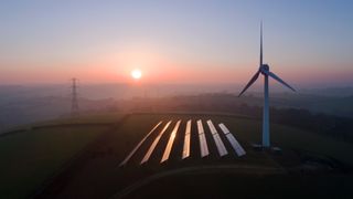 A field of solar panels next to a wind turbine at sunset. 