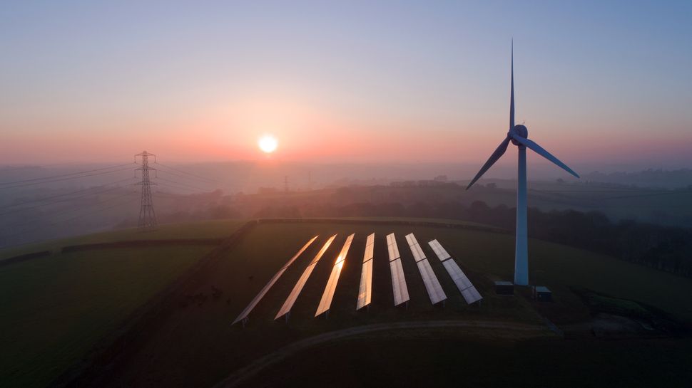 Um campo de painéis solares ao lado de uma turbina eólica ao pôr do sol.