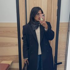 Chiara wears a long black coat, white sweater, jeans, and neckerchief in a mirror selfie 