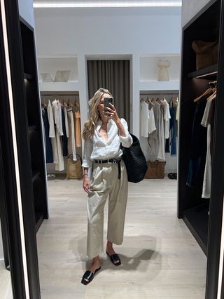 Eliza Huber in the dressing room at Banana Republic wearing a white linen button-down shirt with tan khaki pants, black slides, a black raffia oversize bag, and a black belt.