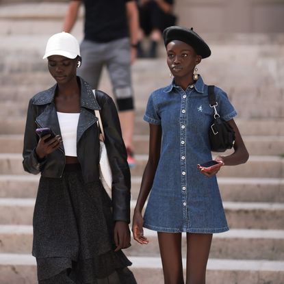 Two women walking down the steps outside a show at Paris fashion week, one is wearing a black skirt, black leather jacket and a white cap, the other is wearing a denim mini dress and a black beret