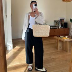 Emily wears white tank and cardigan over dark trousers and slide sandals while carrying a straw tote bag