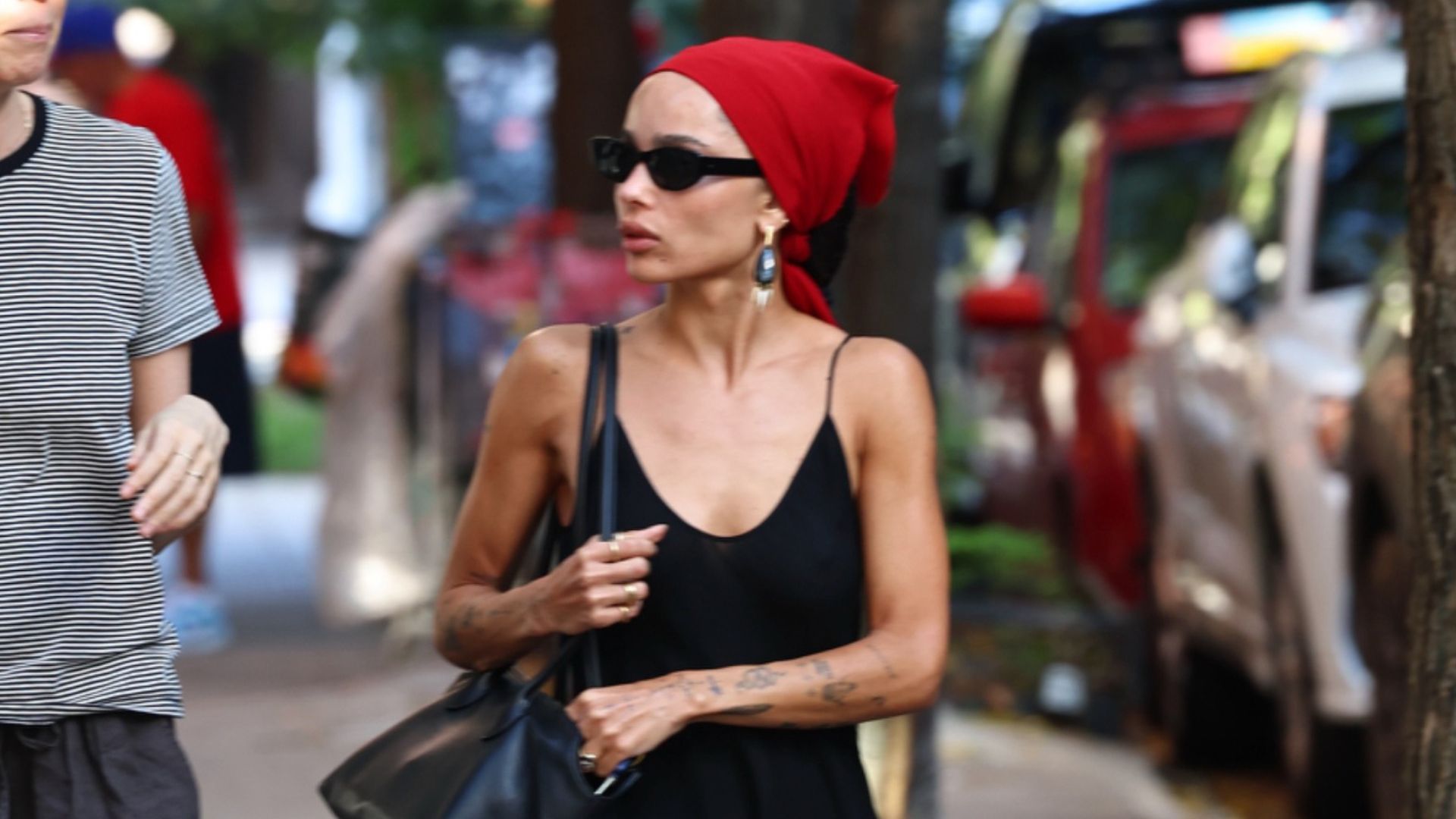 Zoe Kravitz walks down the street wearing a black slip dress with a red bandana, black flip-flops, a black leather bag and oversized earrings.