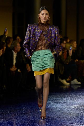 A model at Saint Laurent on the runway during the spring/summer 2025 season wearing a purple brocade jacket with a green layered miniskirt.