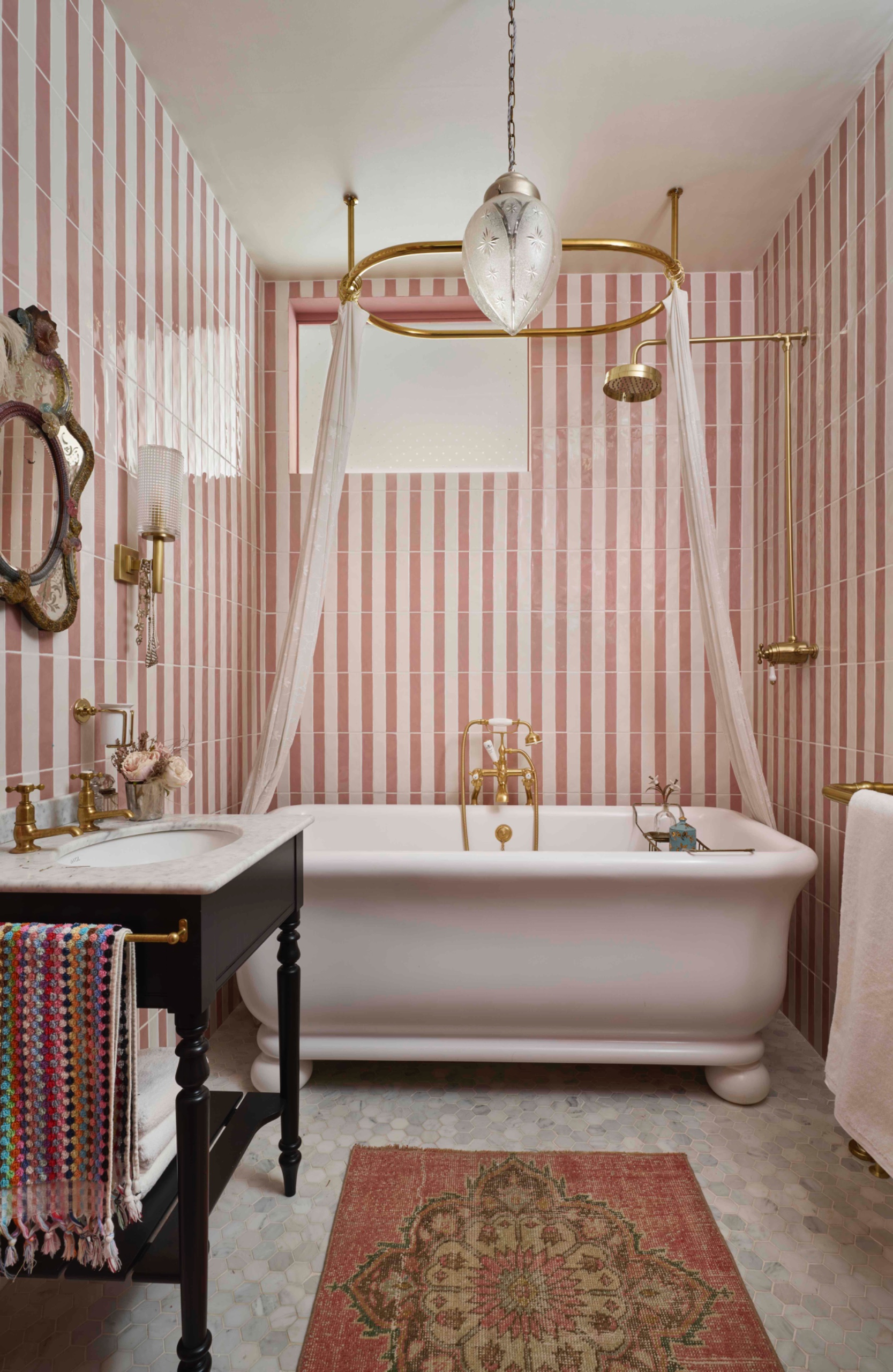 Small bathroom with pink and white vertical tile stripes, a freestanding white bath, brass shower fittings, sheer curtains and a dark vanity.