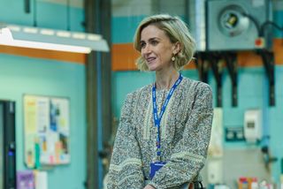 George (Katherine Kelly) stands in the ambulance depot's main office, wearing a floral peasant blouse and a staff lanyard
