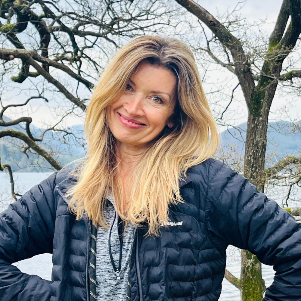 female with long blond hair wearing navy puffer jacket and grey top stood outside with trees and lake in background