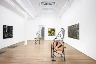 A series of artworks, including two human-shaped chairs, sit in a lit gallery with wooden floors and white walls.