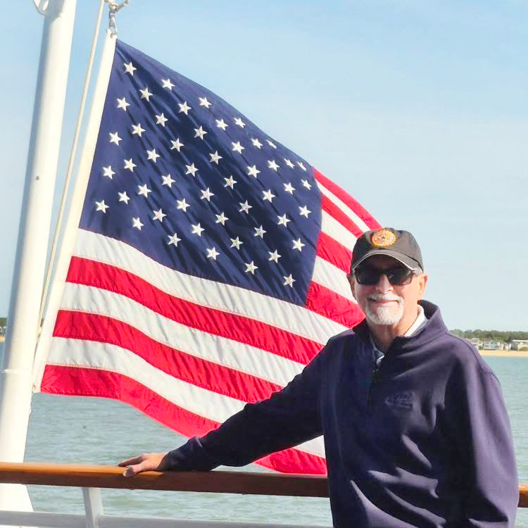 Don Ambrose smiling in front of an American flag on the rail of a boat.