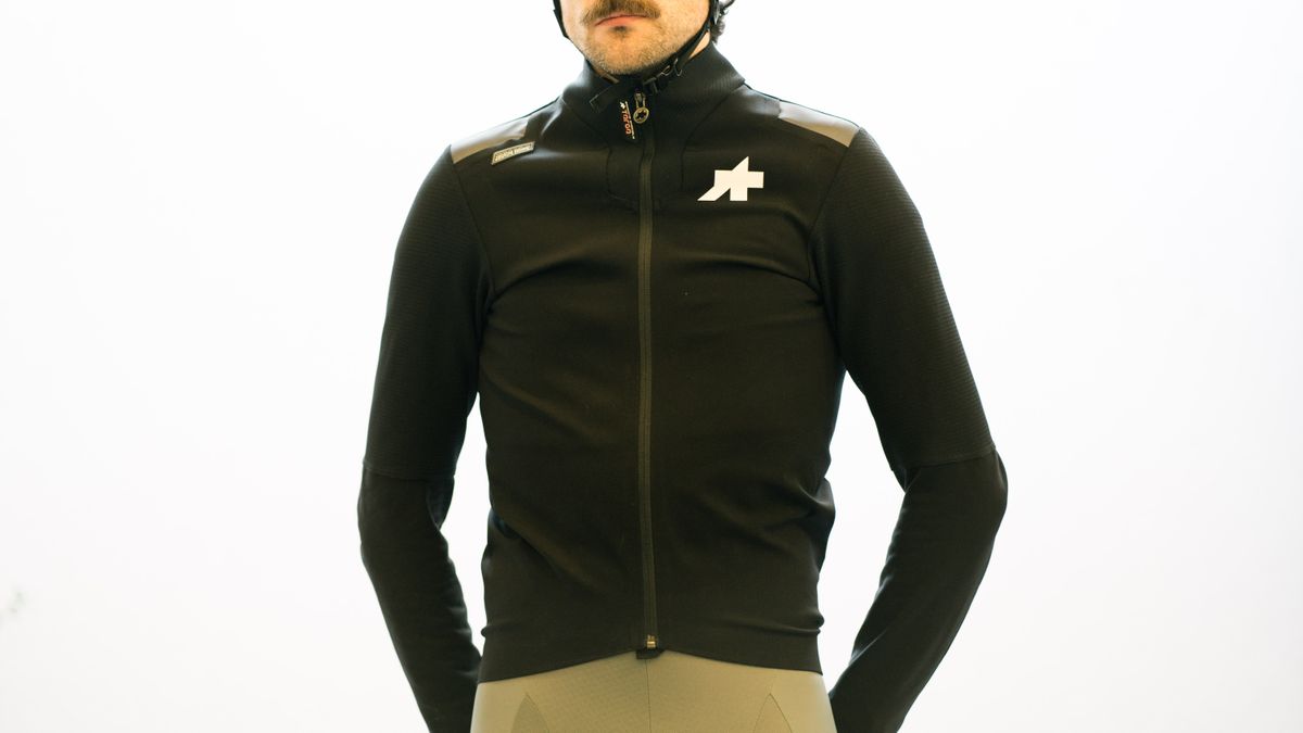 Will stands against a white wall while wearing the black Assos Johdah cycling jacket