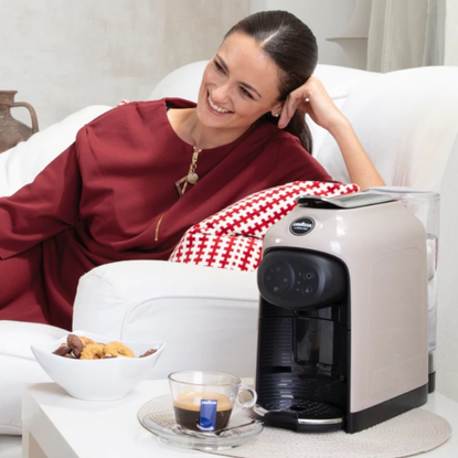 Woman sitting on couch with the Lavazza Idola Capsule coffee machine next to her on a table