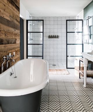 bathroom with reclaimed wood panelling on wall behind freestanding bath, shower enclosure at end of room, black and white geometric tiled flooring