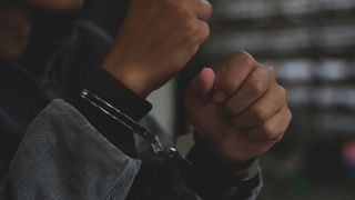 A person is wearing a grey jacket with black cuffs. They have their hands raised and handcuffs are visible around their wrists.
