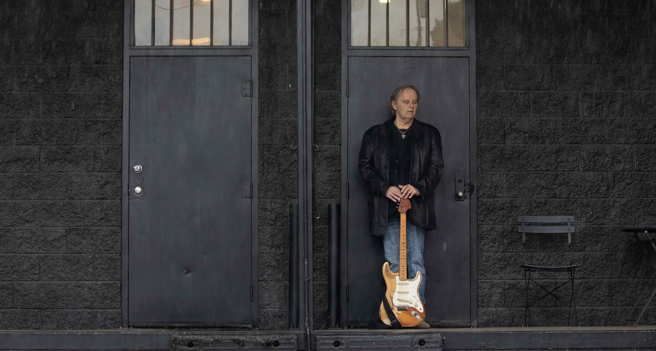 Walter Trout is photographed in front of one of two doors with his hands resting on the headstock of his weather-beaten Stratocaster.