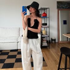 Mimi Nguyen wearing a black cami top, white linen trousers, beaded bag, black raffia bucket hat and blue iPhone case, taking a selfie at home.