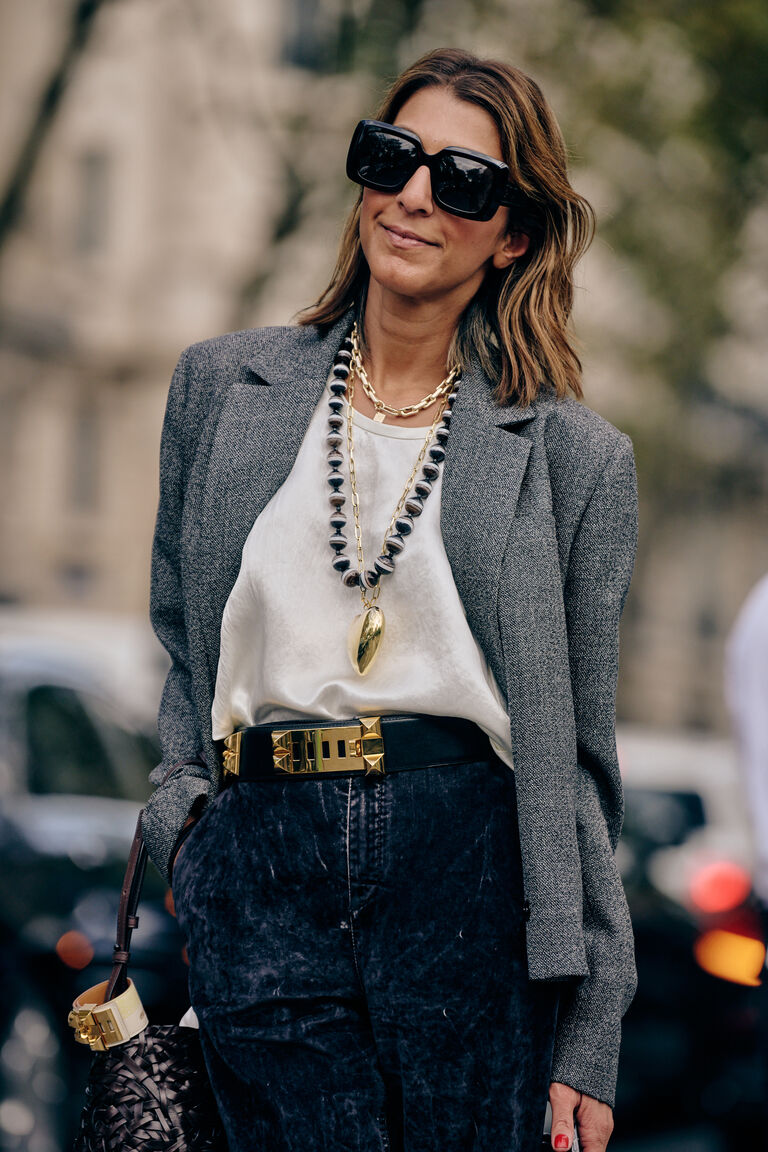 a woman at paris fashion week wearing a white tee shirt, grey blazer, denim jeans, a gray and black beaded necklace, and black and gold belt.