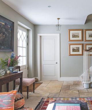 A bright and colorful entryway with tiled flooring, grey walls, patterned rug and cushions, wooden console table with flowers and framed wall art with white staircase and white painted door