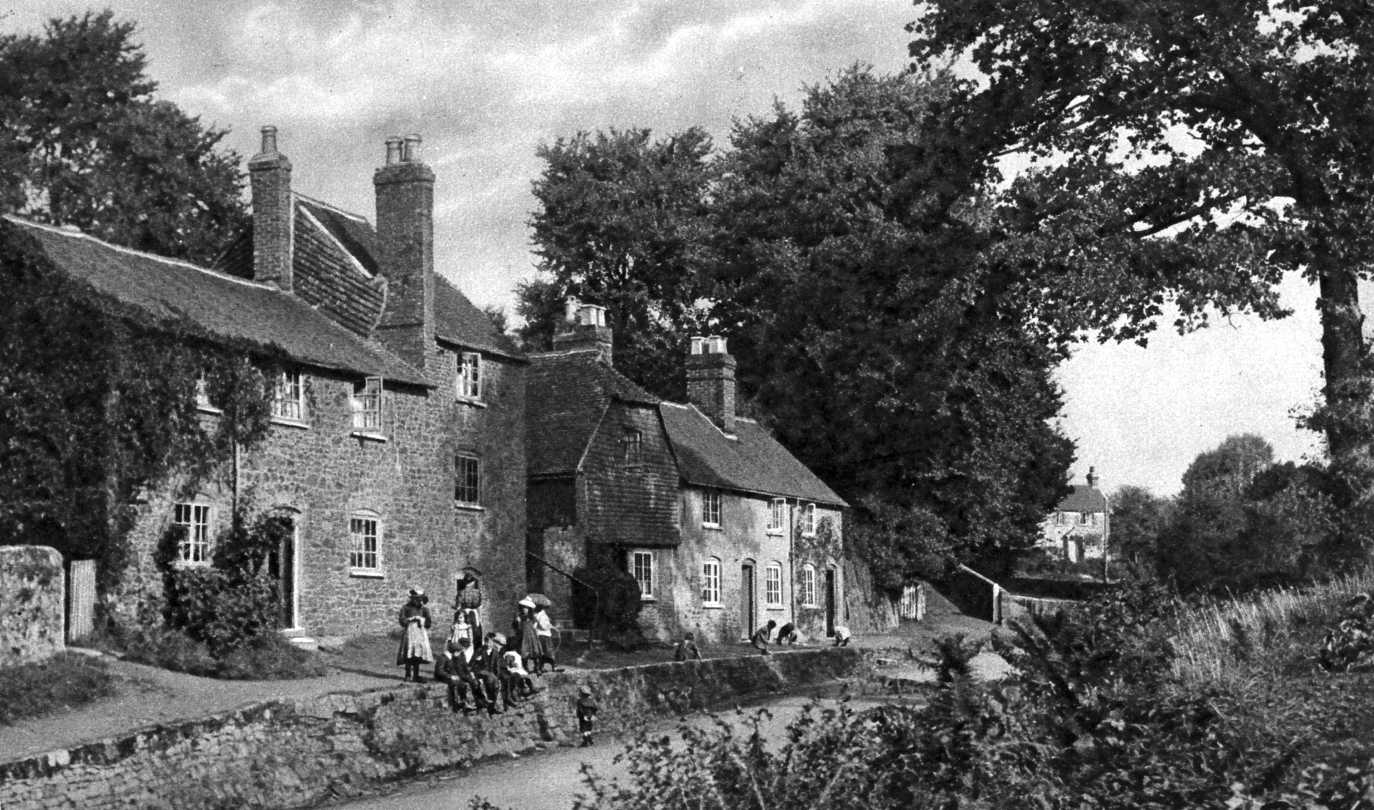 A black and white photograph of an old stone house on a river bank. A group of people in 1920s clothing are sitting on its banks