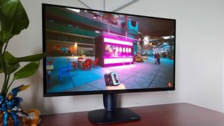 Alienware AW2725Q gaming monitor with Cyberpunk 2077 gameplay on screen featuring player character holding gun looking at neon lit ramen stand.