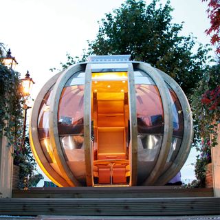 an orange tinted garden room pod with seating and a table inside, sat on wooden decking with draping plants scattered around the outside of it