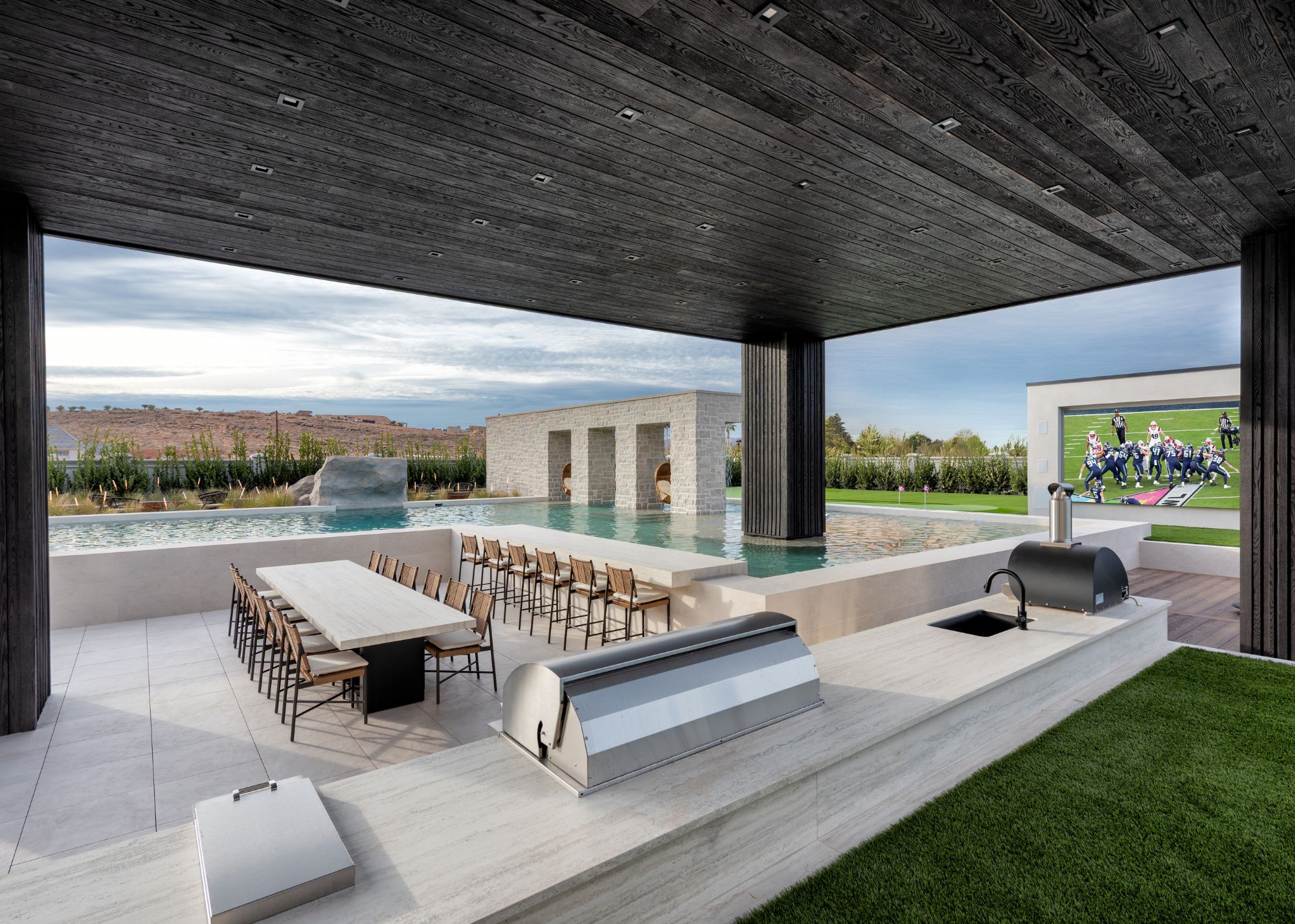 A grand outdoor kitchen and seating area with a huge outdoor tv with american football on the screen and a pool with a black-clad ceiling above the kitchen and seating area