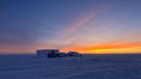 A lone square building with small structures sitting a vast, flat area of snow during twilight.