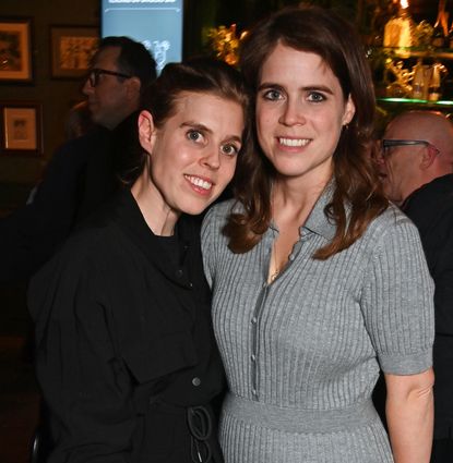 Princess Beatrice wearing black and Princess Eugenie in a gray dress posing in a restaurant