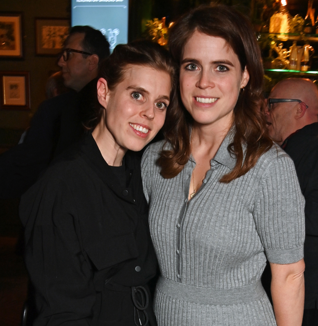 Princess Beatrice wearing black and Princess Eugenie in a gray dress posing in a restaurant
