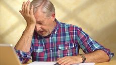 An older man sitting in front of his laptop puts his head in his hand as if he's made a mistake.