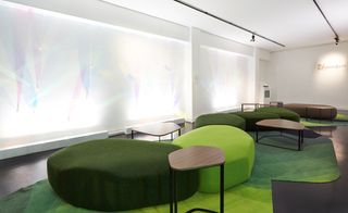A lounge is furnished with light and dark green seating modules in asymmetrical shapes, complete with a carpet in different shades of green and asymmetrical wood and metal tables.
