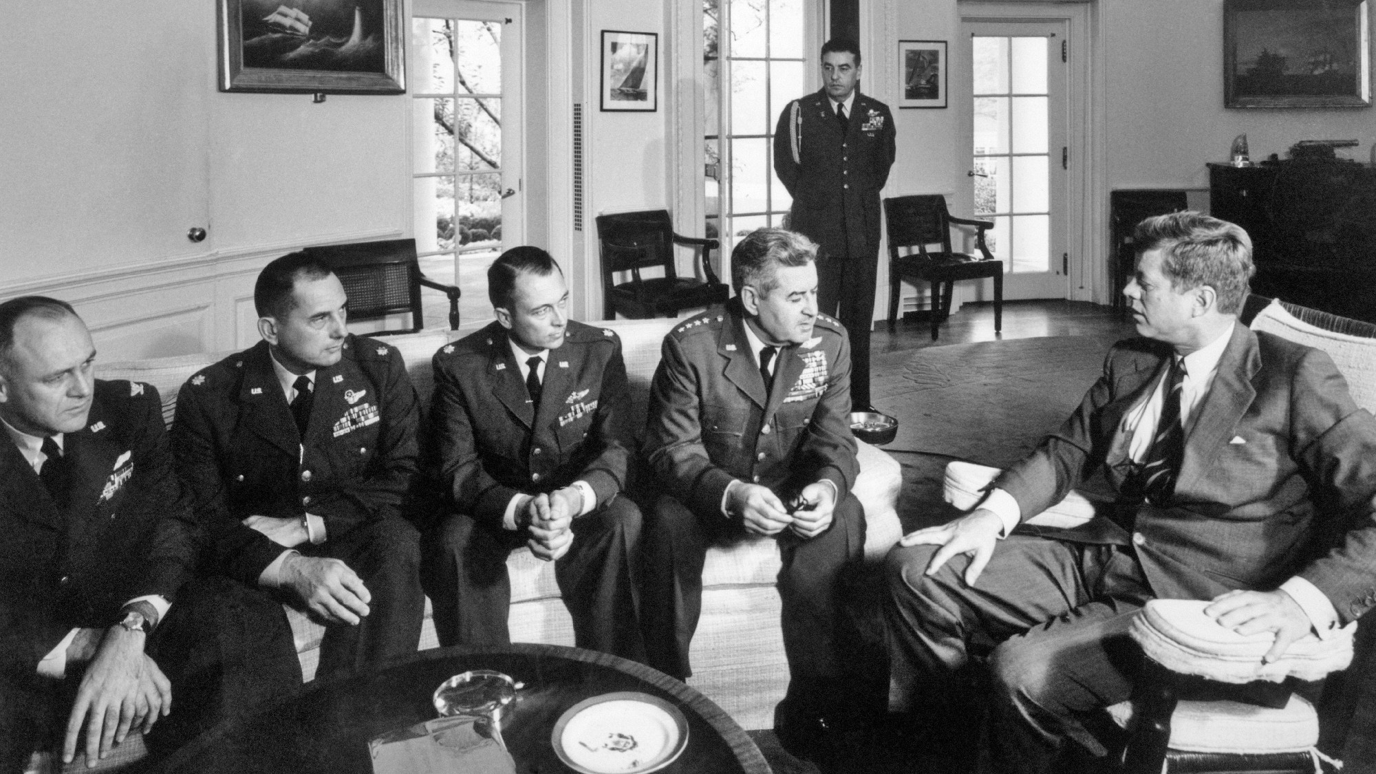 President John F Kennedy pictured alongside men in military uniform, in the Oval Office, during the Cuban Missile Crisis
