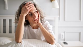 A woman with short gray wavy hair wearing a white t-shirt holds her forehead in one hand, her elbows propped up on a pillow on her bed. 