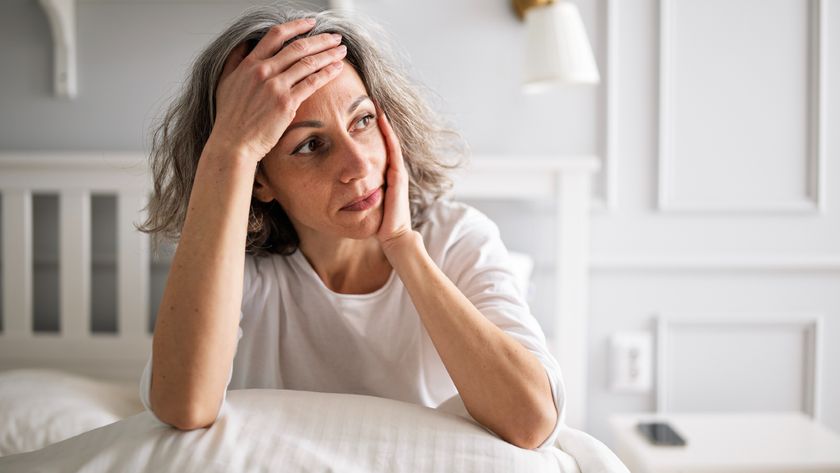 A woman with short gray wavy hair wearing a white t-shirt holds her forehead in one hand, her elbows propped up on a pillow on her bed. 