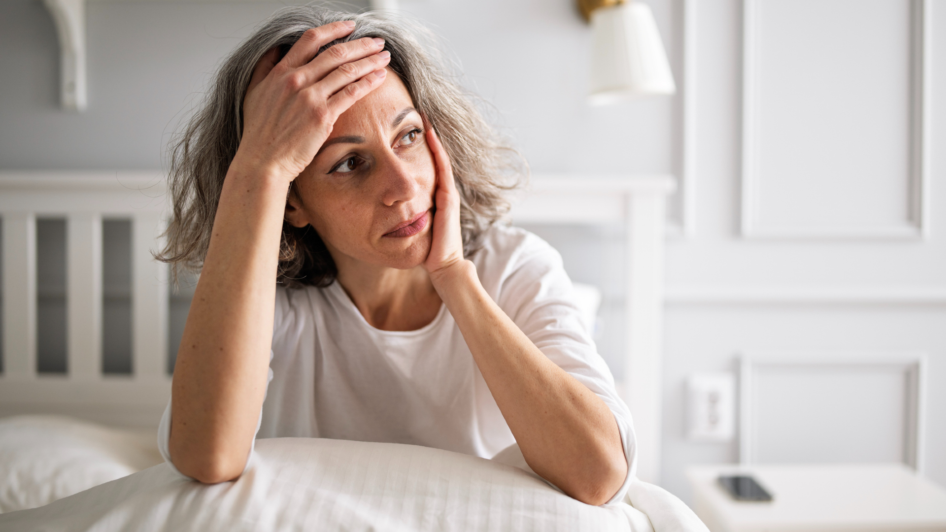 A woman with short gray wavy hair wearing a white t-shirt holds her forehead in one hand, her elbows propped up on a pillow on her bed. 