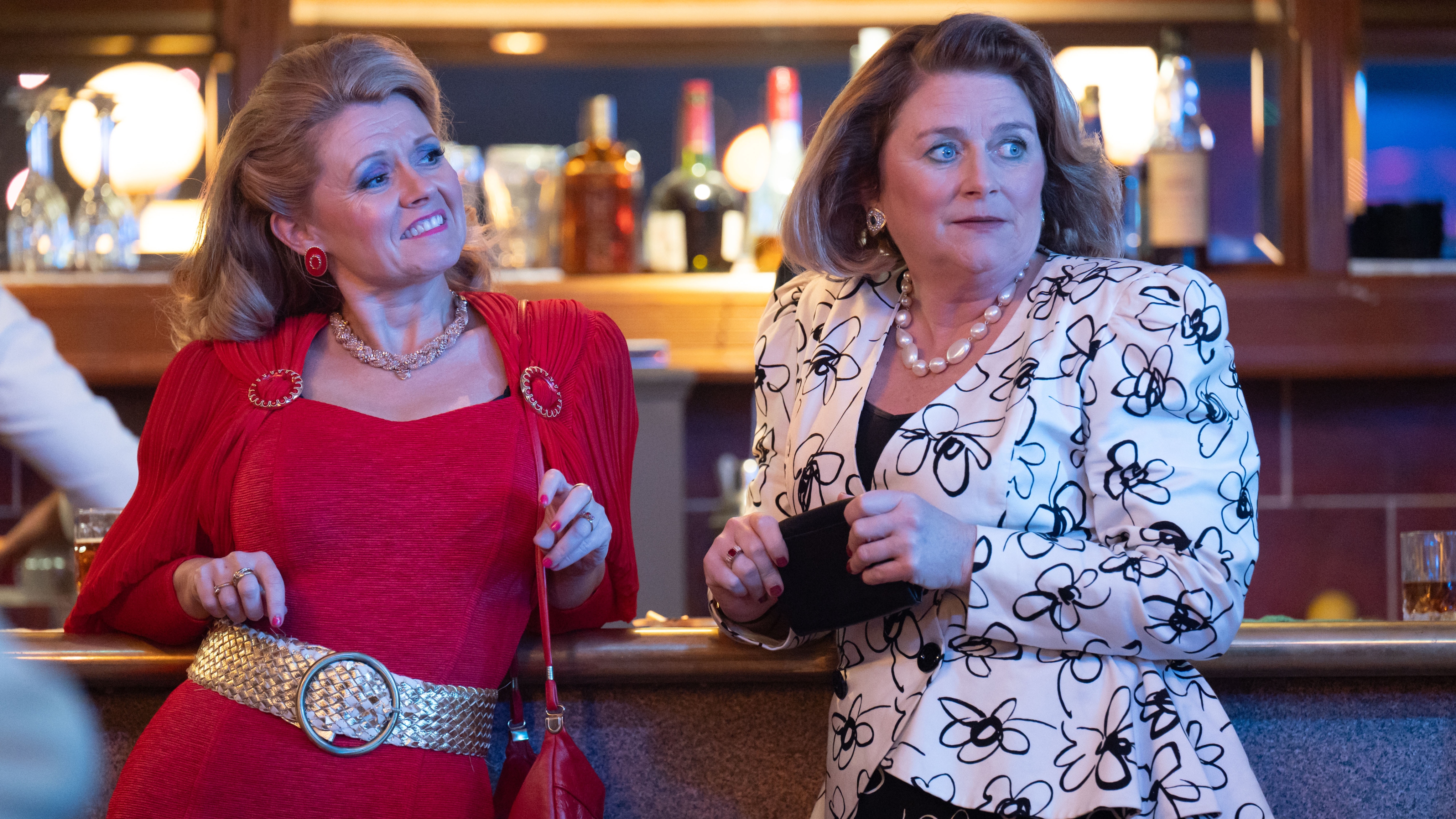 Kath (Sian Gibson) and Diane (Rosie Cavaliero) are standing with their backs to the bar at a posh function. Kath is wearing a tight red dress with a statement gold belt, and Diane is in a white blazer with a black floral pattern. Kath looks like she's enjoying herself, while Diane looks a little nervous.
