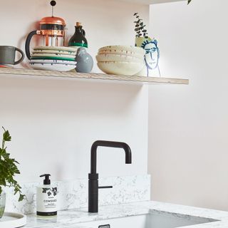 A white kitchen basin with matte- black tap, and wooden shelf with assorted ceramicware including bowls and cafetiere on display
