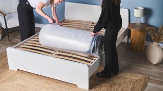 Two testers unboxing a memory foam mattress that's rolled in plastic onto a bed frame