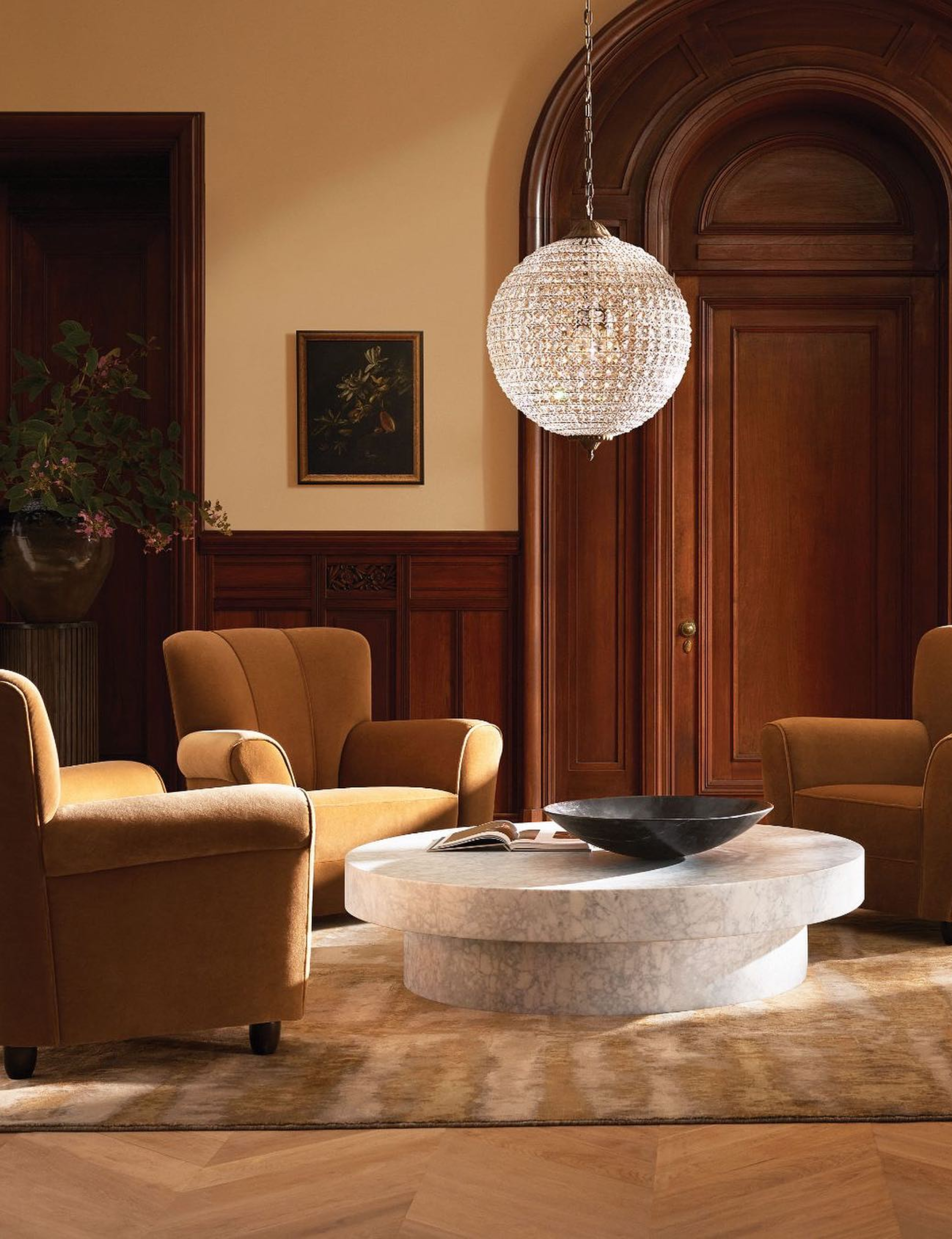 Swanky living room with herringbone wooden floors and decorative paneling decorated with a glittering crystal-cut chandelier, suede armchairs, and an oversized marble coffee table