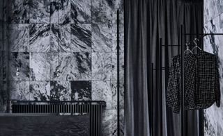 Dark gray marble tiled wall with a black curtain to the right. In front of it is a clothes rack with shirts hung.