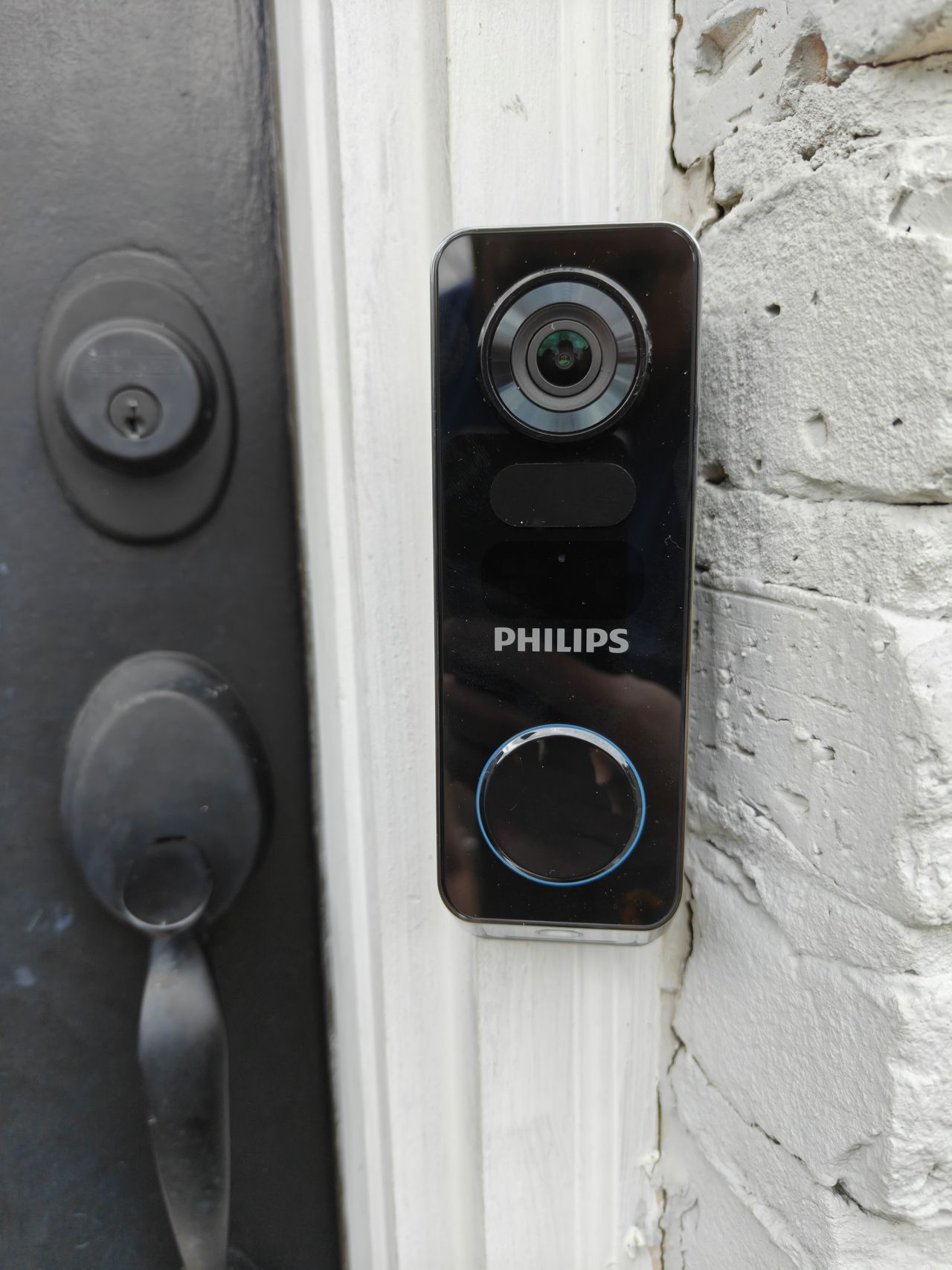 Philips 7000 Series 2K Video Doorbell mounted on white doorframe next to gray door and white bricks.