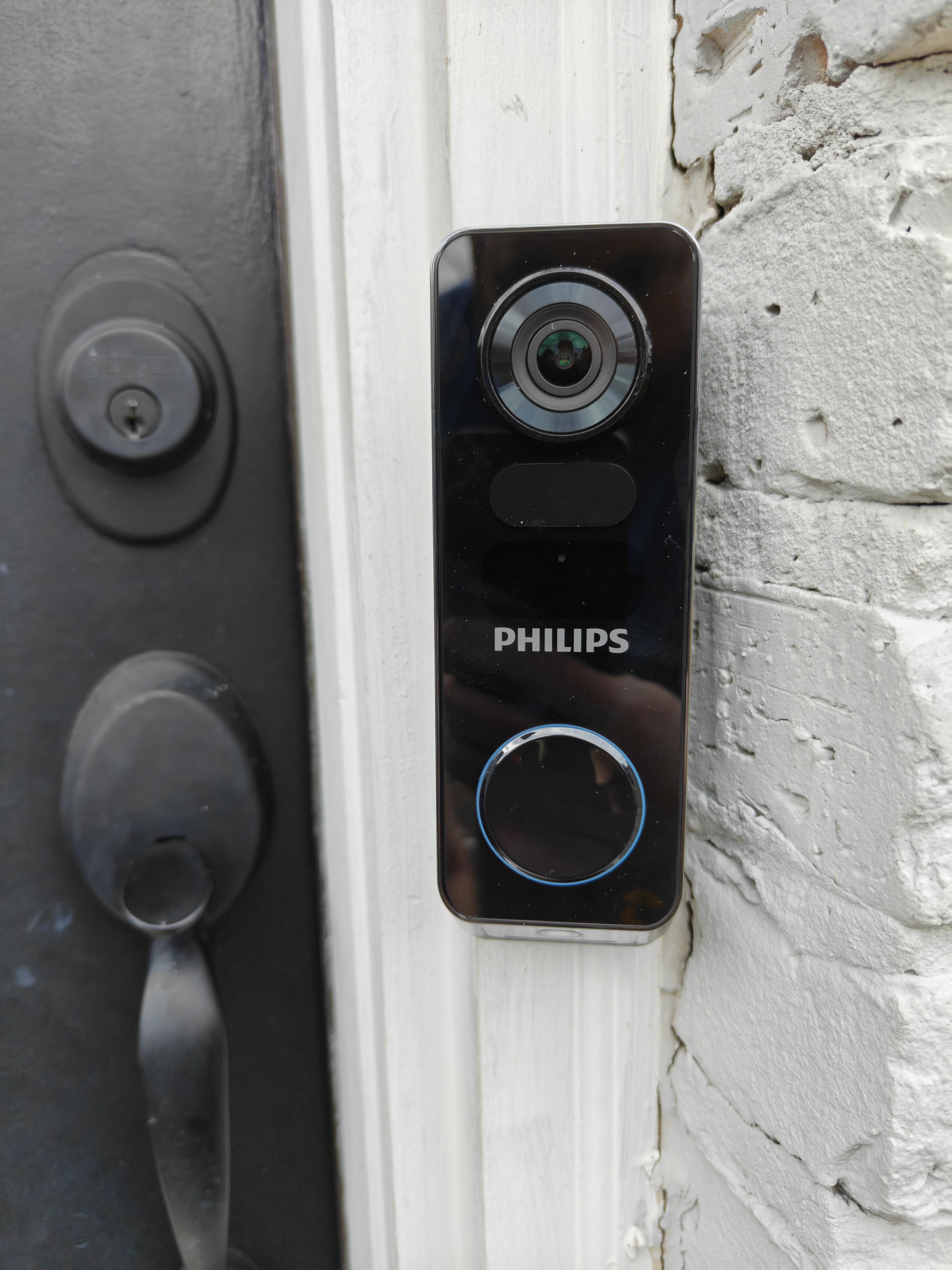 Philips 7000 Series Video Doorbell mounted next to gray front door with white bricks pictured to the right.