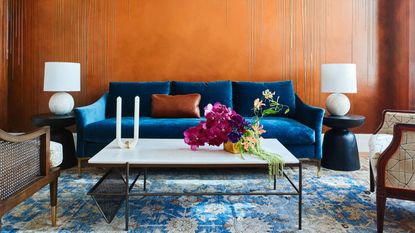 living room with orange wall, blue sofa, patterned rug, marble coffee table, two side tables with white lamp on them, and two cane armchairs