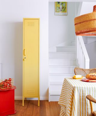 White and yellow kitchen with vertical locker style storage in yellow, yellow and white striped tablecloth, rattan chairs and pendant light, artwork at top of white painted stairs
