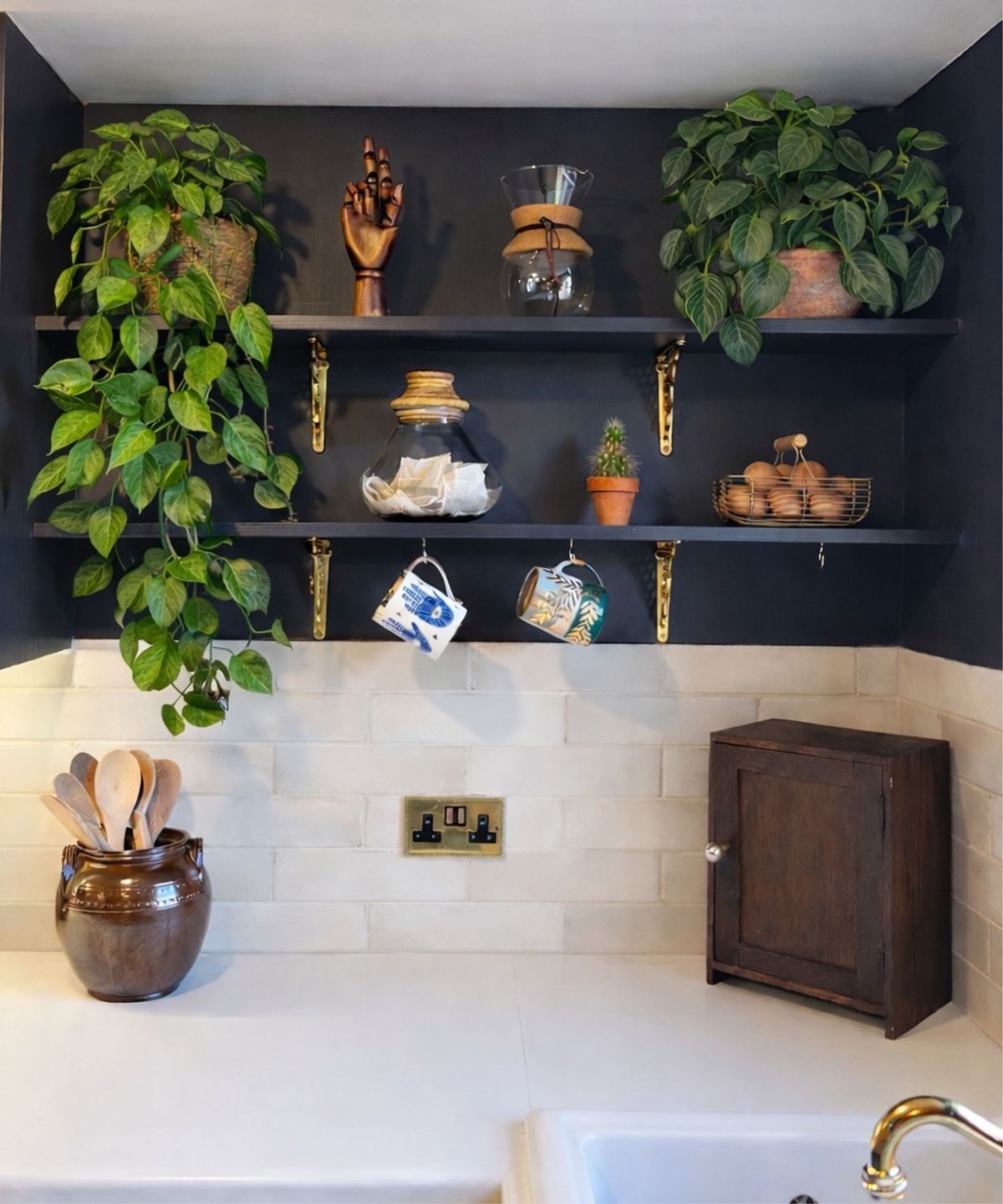 Navy kitchen shelves with lush trailing plants, brass brackets, glass jar, mugs, cactus and eggs, above white tiles and counter with wooden utensils and small cabinet.