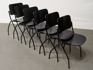 Six black chairs staked in a row and photographed in a room with white walls and grey floor