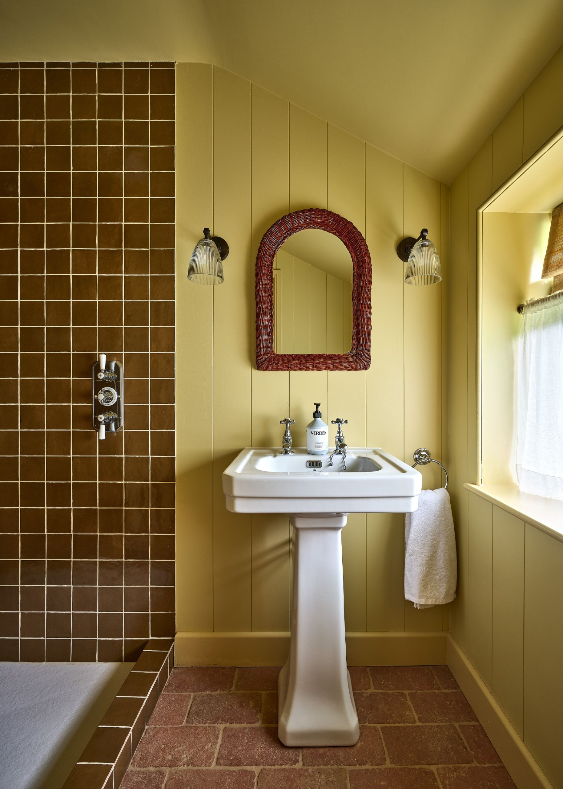 A yellow color-drenched bathroom with wall panelling, a white sink, an arched dark brown mirror, and two wall sconces. Terracotta-tiled flooring and caramel square wall tiles in the shower.