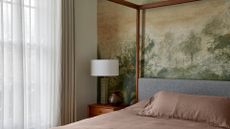 A calming bedroom with a nature mural, a green table lamp, a vase, taupe bedding, and sheer curtains