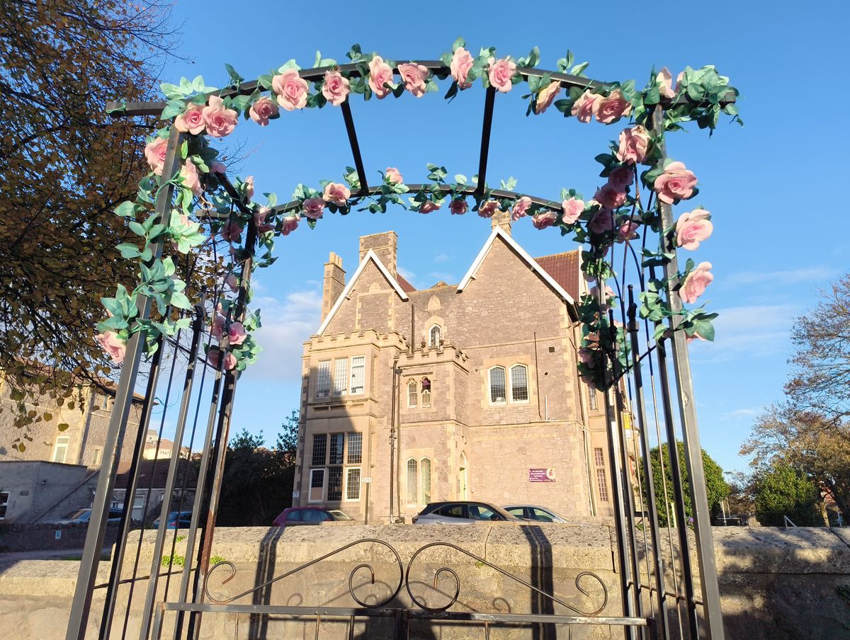 A black metal garden arch adorned with fake pink roses and greenery frames a view of a large, distinctive stone building with multiple gables and white-framed windows under a bright blue sky.
