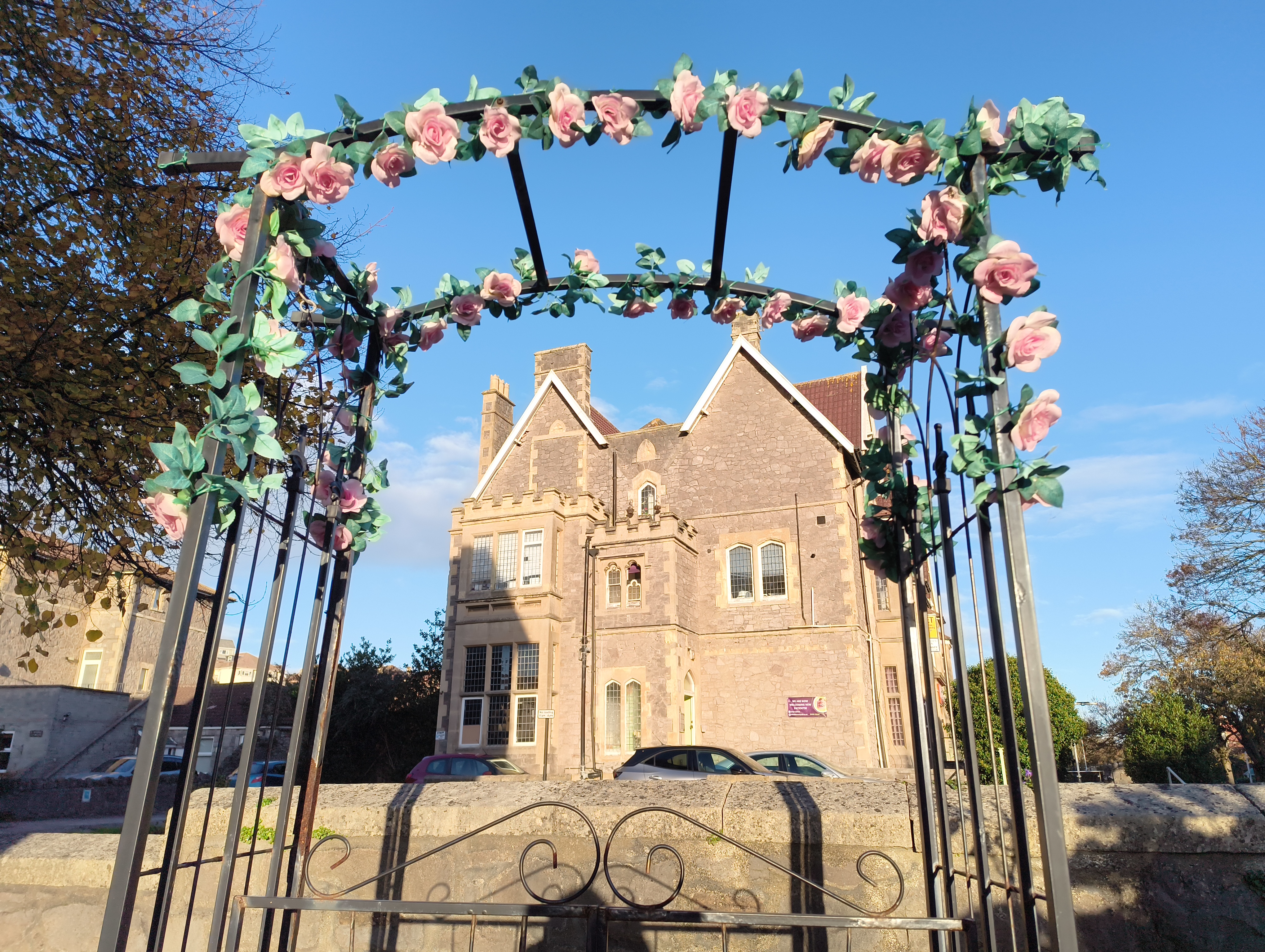 A black metal garden arch adorned with fake pink roses and greenery frames a view of a large, distinctive stone building with multiple gables and white-framed windows under a bright blue sky.