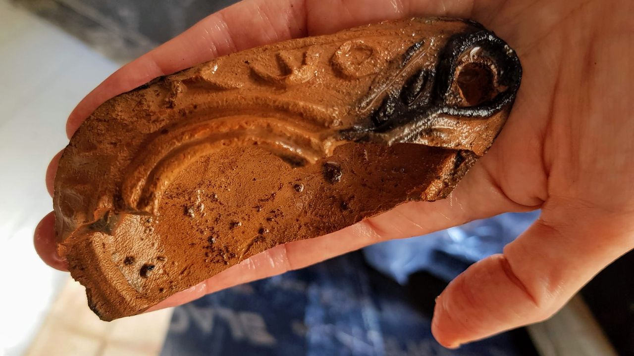 A fragment of brown pottery with some black marks, displayed on someone&rsquo;s palm. It is a piece of oil lamp with a Greek inscription that reads "the light of Christ shines for all."